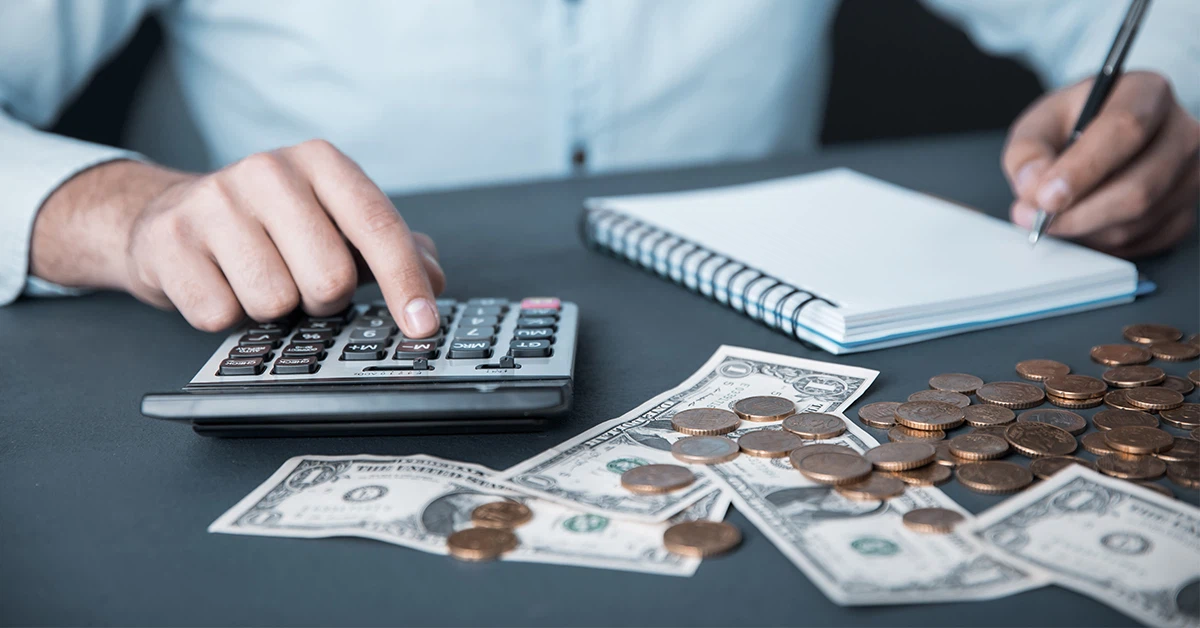 cropped photo of a man making calculations on a calculator; he writes in a notebook; dollar bills and coins are strewn on the table (featured image for article: "The Hidden Costs of In-House Payroll: What Small Businesses Need to Know")