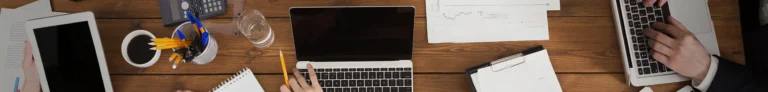 overhead photo of a meeting with laptops, papers, writing utensils, beverages, and hands around a wood table (Green Payroll Employee Login page hero image)