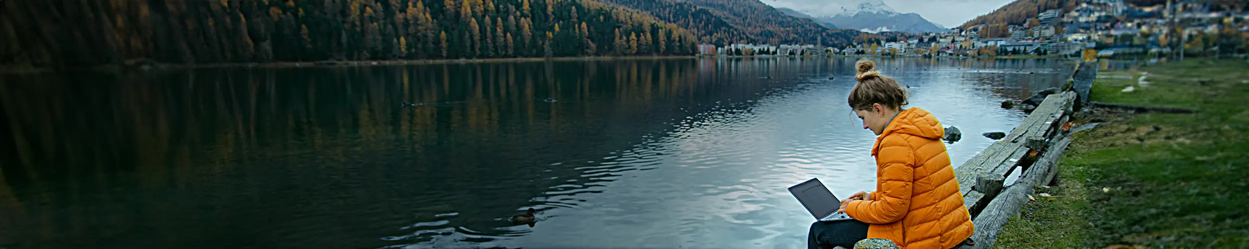 photograph of a woman with a messy bun in an orange puffy jacket sitting next to the water in the mountains, working on a laptop (Green Payroll hero image)