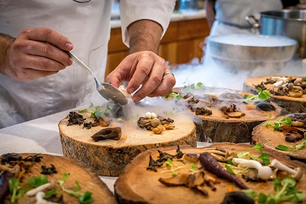 close-up photo of a chef plating a high-end mushroom tasting dish on wood slices as plates with dry ice smoke pouring off of his spoon