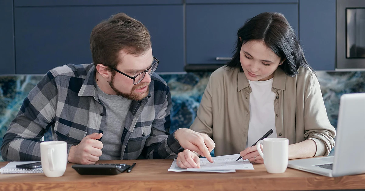 photograph of a man and woman sitting at a table and working together on paperwork with coffee cups, a laptop and a calculator on the table (featured image: "Mastering Payroll Compliance: A Guide for Businesses" article)