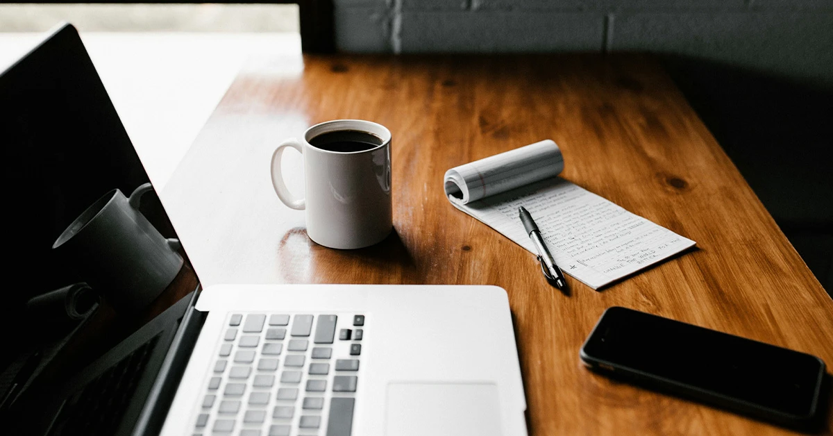 photo of a laptop sitting on a wooden table with a mug of black coffee, a mini legal pad with notes, a pen, and a smartphone (featured image: "HR Compliance Checklist: Essential Steps for Employers" article)