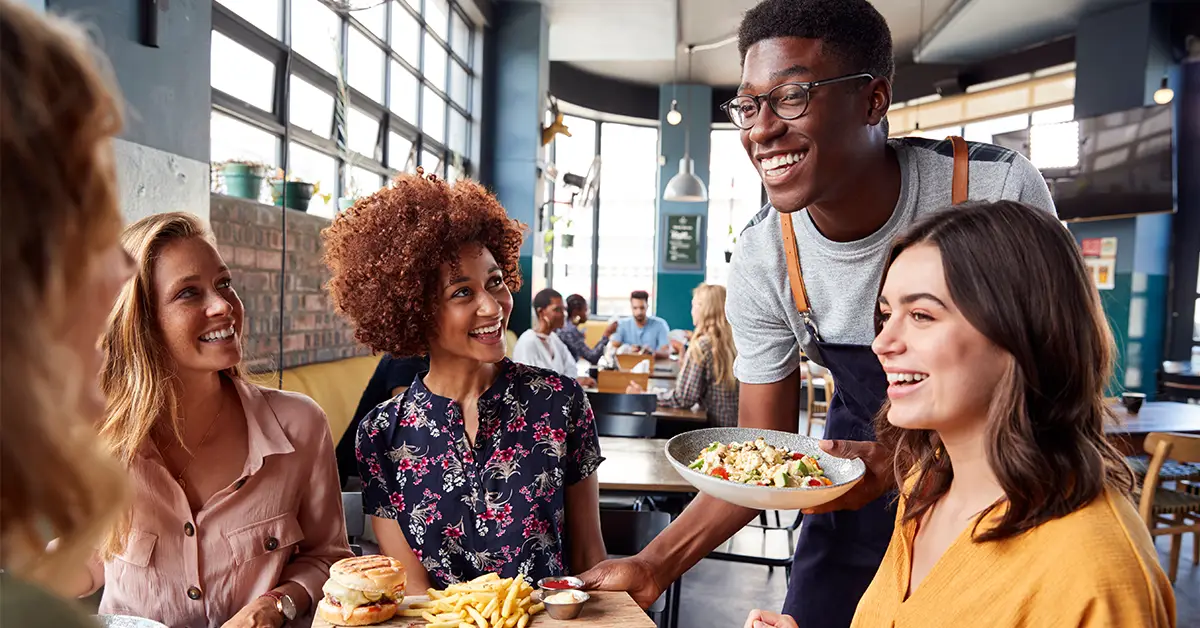 photo of a group of female friends at a trendy restaurant, a server wearing glasses and an apron is smiling at the group and delivering food (featured image: "No Tax on Tips: Guide for Employees and Payroll Teams" article)