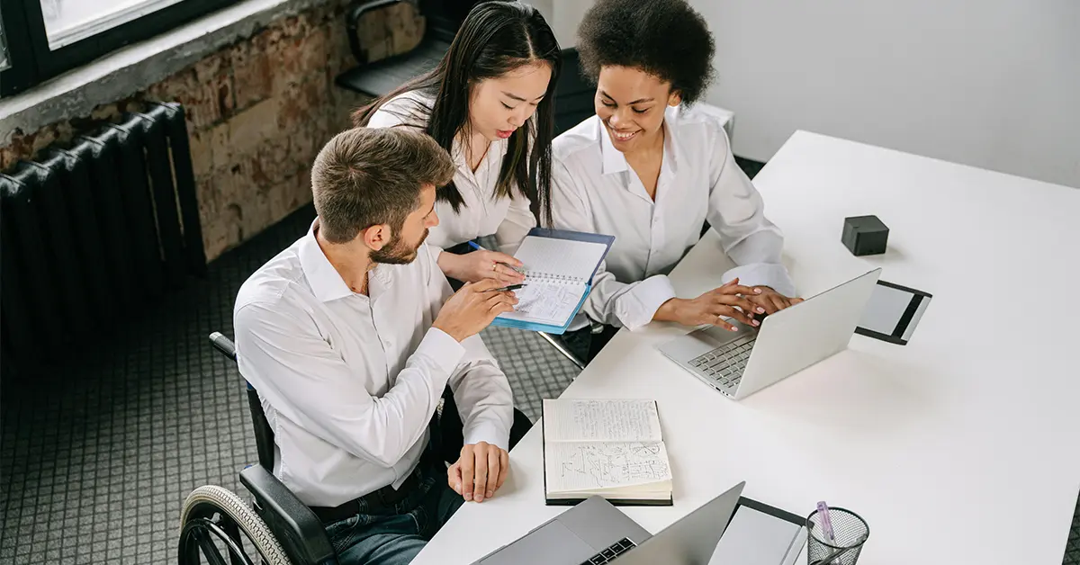 High-angle photo of a diverse group of colleagues working together around a table: a Caucasian man in a wheelchair, an Asian woman, and a Black woman work with laptops and notebooks with notes and graphs. (featured image: "Work Opportunity Tax Credit (WOTC): Eligibility, Process & Value" article)